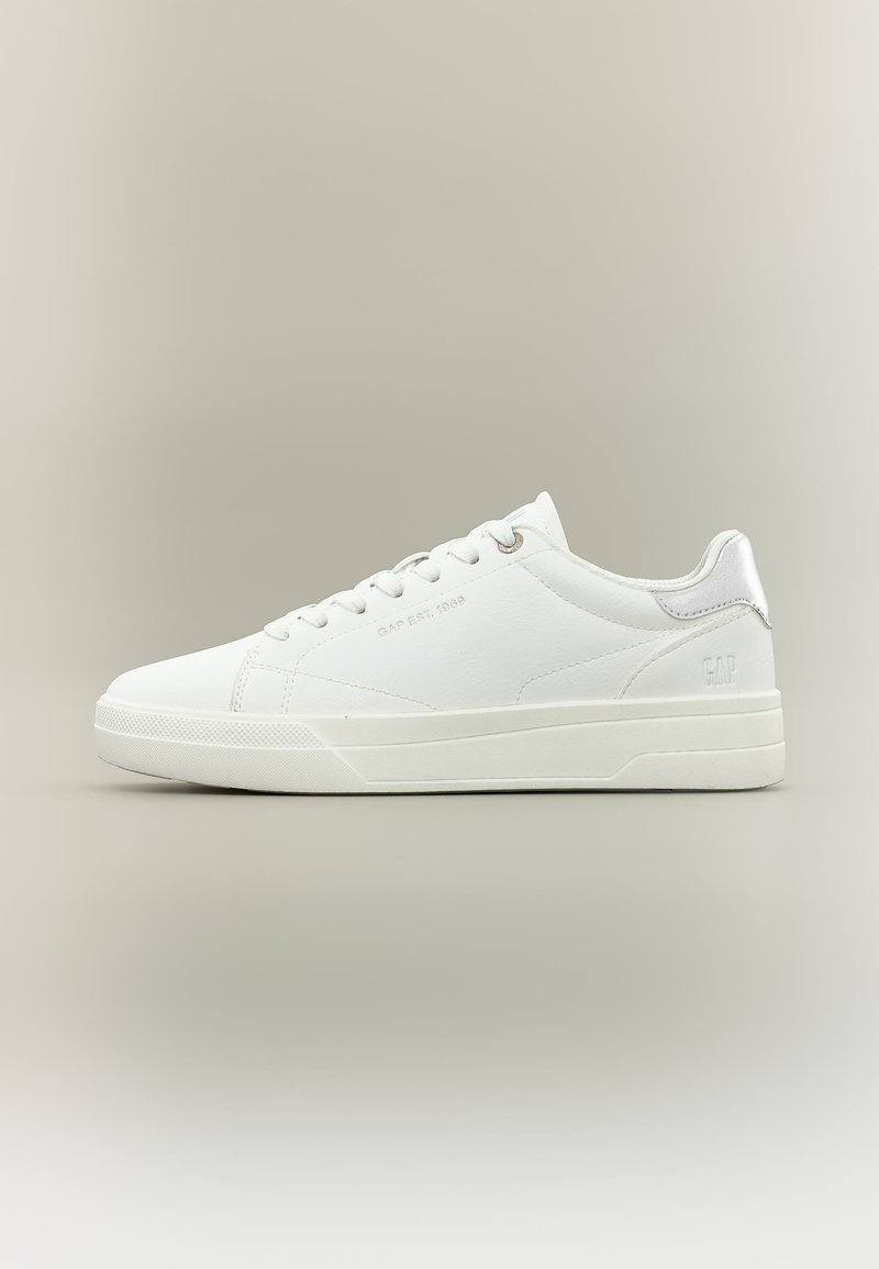 Baskets blanches basses à lacets, avec un branding discret sur le côté et le talon, et une semelle en caoutchouc sur un fond beige neutre.