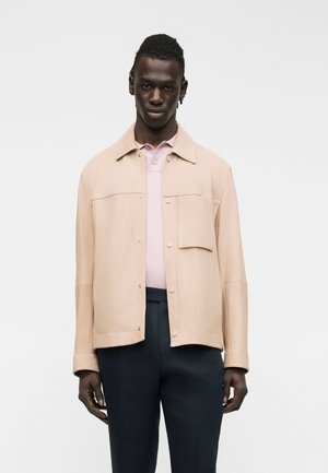 Άνδρας που φοράει μπεζ σακάκι με κουμπιά πάνω από ένα ανοιχτό ροζ πουκάμισο πόλο και μαύρο παντελόνι, στέκεται μπροστά σε απλό λευκό φόντο.