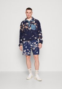 Marineblå fleece-pullover med flamingo-mønster, lyseblå lomme og krage. Matchende shorts har et lignende print. Hvite joggesko og sokker.