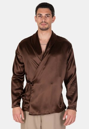 Uomo che indossa una camicia lunga marrone in raso stile avvolgente, annodata di lato, abbinata a pantaloni beige, in piedi davanti a uno sfondo semplice.