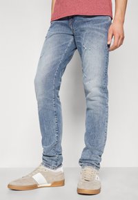 Ljusblå denimjeans med slim fit, med slitna områden och en urblekt finish. Parat med beige sneakers och en röd topp.