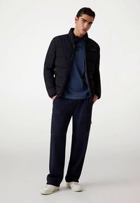 Jachetă matlasată neagră peste un top albastru închis, asortată cu pantaloni închiși la culoare cu buzunare laterale și adidași albi.