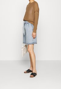 Maglione lavorato a maglia marrone chiaro, pantaloncini in denim con vita annodata, sandali neri a scivolo e una borsa intrecciata; su uno sfondo bianco.