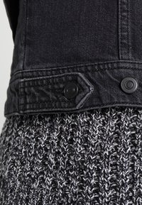 Gros plan de l'ourlet d'une veste en denim noire avec des boutons-pression noirs sur un tissu tricoté noir et blanc texturé.