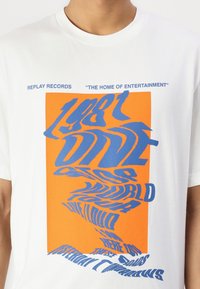 Balta medvilninė marškinėliai su ryškiu iškraipytu mėlynu tekstu ant oranžinio fono. Tekste yra "Replay Records" ir "1981 Dive" su papildomu žodynu.