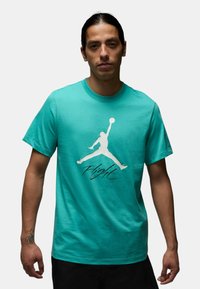 T-shirt in cotone turchese con grande logo Jumpman bianco e la parola "Flight" in corsivo, maniche corte e collo girocollo.