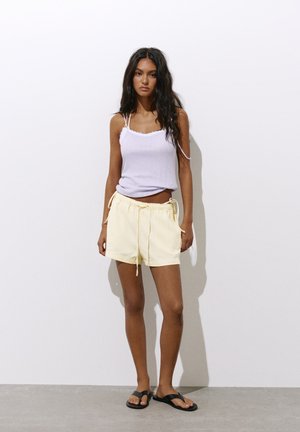 Junge Frau mit langen dunklen Haaren, die ein weißes Spaghettiträger-Top, hellgelbe Shorts und schwarze Sandalen trägt und an einer weißen Wand steht.