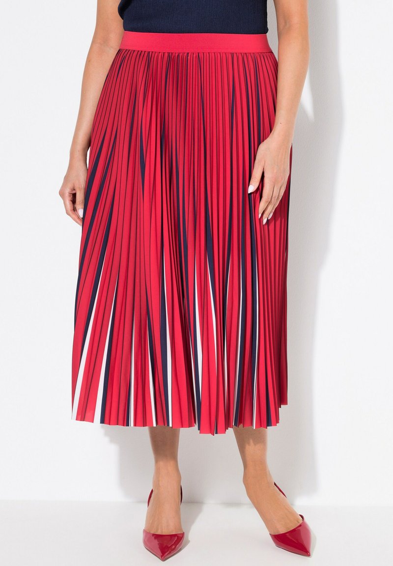Jupe plissée rouge avec des rayures bleu marine et blanches, en tissu texturé, longueur mi-mollet, associée à des escarpins rouges à talons pointus. Ceinture élastique visible.