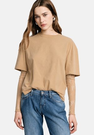Donna con lunghi capelli castani indossa una t-shirt beige oversize sopra maniche lunghe in pizzo trasparente e jeans blu larghi, in piedi con mani rilassate.