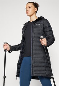 Zwarte pufferjas met capuchon, voorzien van een horizontaal quiltpatroon, een rits aan de voorkant en het Columbia-logo, gedragen met blauwe leggings en trekkingstokken.