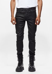 Hombre con pantalones de cuero negros ajustados y botas de combate negras con cordones, de pie contra un fondo blanco.