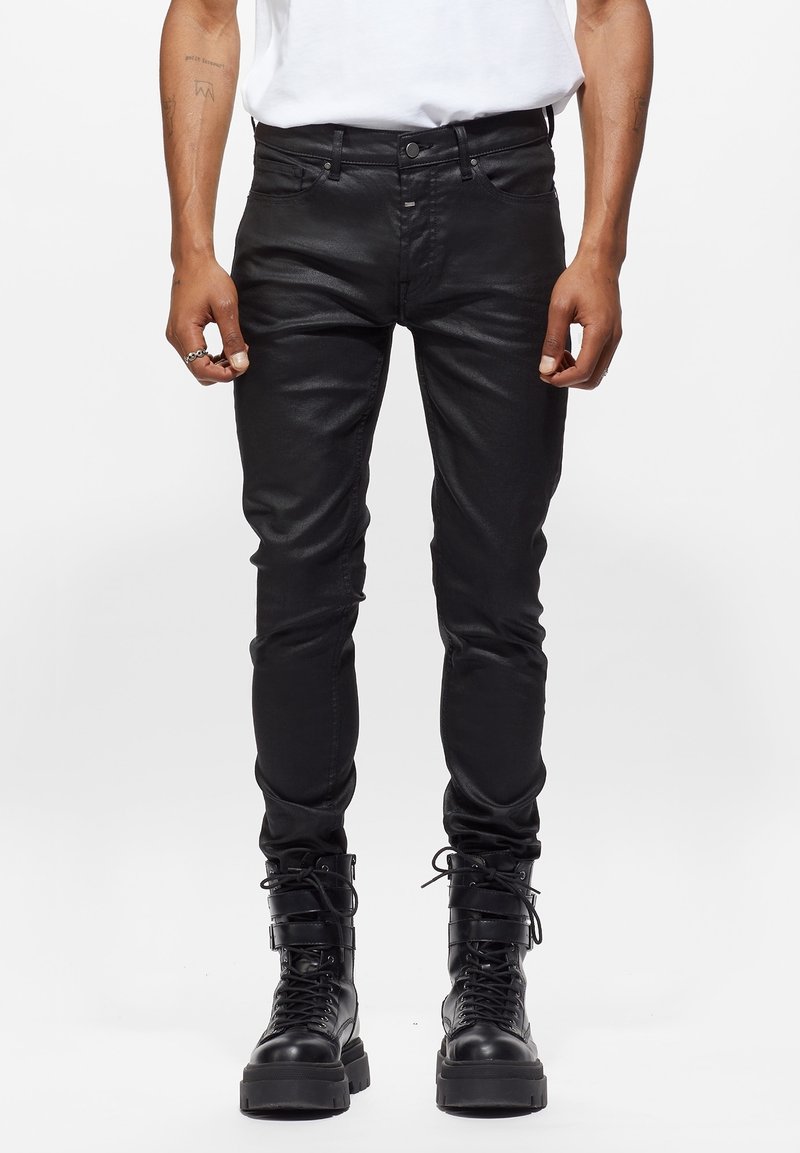 Hombre con pantalones de cuero negros ajustados y botas de combate negras con cordones, de pie contra un fondo blanco.