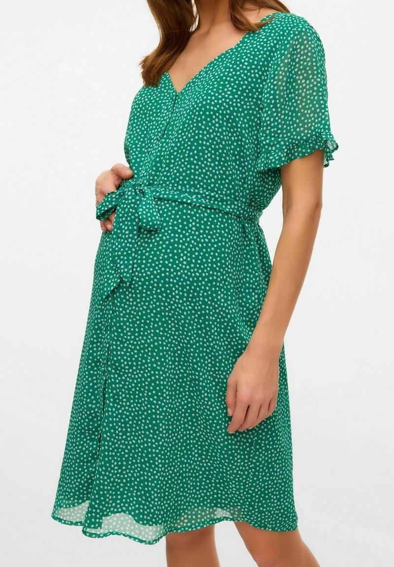 Vestido verde de lunares, hecho de una tela ligera, con mangas cortas de volante, escote en V y un cinturón atado en la cintura.