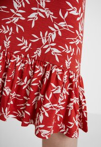 Röd klänning med vitt lövmönster, med en utsvängd volangkant. Tillverkad av mjukt tyg, har den en figursydd bodice och resårmidja.