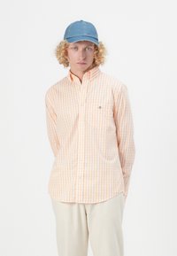 Camicia a maniche corte, con bottoni, in un motivo a quadretti arancione chiaro e bianco, dotata di una tasca sul petto e di un logo discreto. Abbinata a un cappellino in denim.