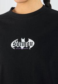 Black cotton t-shirt featuring a white and purple Batman logo with the text "OUT OF THE SHADOWS" in stylized font, round neckline.