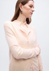 Ελαφρύ κάρντιγκαν σε χρώμα ροδακινί, κατασκευασμένο από μαλακό υλικό, με κουμπιά εμπρός, γιακά και μακριά μανίκια, με απαλή υφή και διακριτικό γκλίτερ.