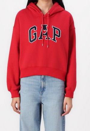 Person mit rotem GAP-Hoodie mit Frontlogo und hellblauer Jeans, steht vor einem schlichten weißen Hintergrund.