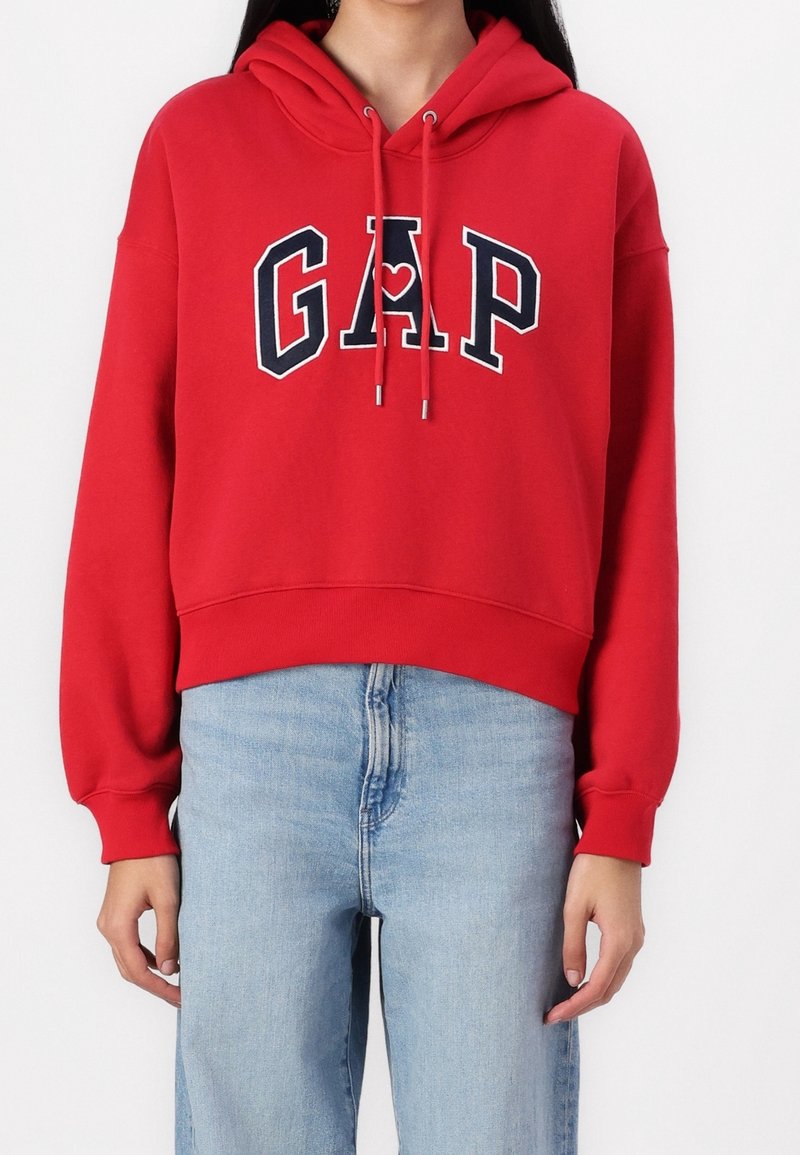 Personne portant un sweat à capuche GAP rouge avec un logo à l’avant et un jean bleu clair, debout sur un fond blanc uni.