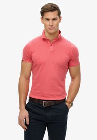 Polo in colore corallo con colletto, maniche corte e una texture sottile. Abbinato a pantaloni scuri e una cintura marrone.