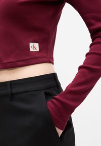 Top scurt burgund cu dungi, cu mâneci lungi, asortați cu pantaloni negri cu talie înaltă, care au un buzunar. Pachet cu logo pe marginea topului.