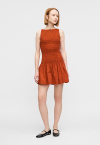 Κοντό, αμάνικο φόρεμα σε χρώμα σκουριάς με μπούστο με σμάλτο και φουσκωτή βάση. Φοριέται με μαύρα φλατ παπούτσια με διακοσμητικά στοιχεία.