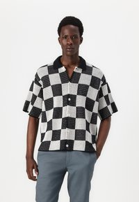 Chemise à manches courtes avec un motif à damier noir et blanc, en tissu tricoté texturé, et un col noir uni.