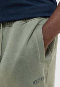 Osoba o ciemnej skórze, ubrana w jasnooliwkowe spodnie dresowe, z ręką w kieszeni i logo "MUSTANG" obok kieszeni.