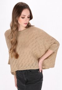 Pulover bej tricotat cu un model deschis din dantelă, mâneci scurte, largi și o croială relaxată, asortat cu blugi negri.
