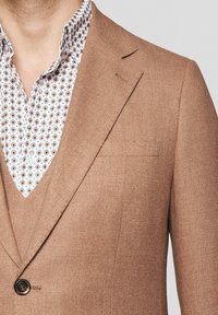Jasnobrązowy żakiet wykonany z teksturowanej tkaniny, z dopasowanym krojem, klapami notch i pojedynczym zapięciem na guzik. Pod spodem znajduje się biała koszula w wzory.