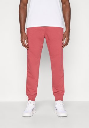Rode sweatpants van een zacht, rekbaar materiaal. Strakke boorden bij de enkels. Draagt een eenvoudig wit T-shirt en witte sportschoenen.