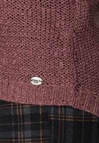 Strickpullover in weinrot mit strukturiertem Muster; enthält ein silbernes Rundschild mit "ONLY." Marke. Kombiniert mit einem schwarz-braunen karonierten Unterteil.