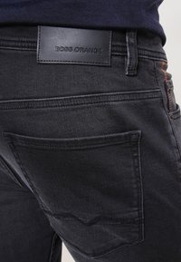 Vaqueros de denim negro con un parche de logo en la parte trasera, con un acabado desgastado, bolsillos traseros cosidos con un diseño curvado y costuras en hilo contrastante.