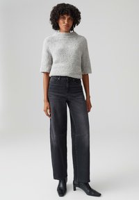 Maglione grigio, morbido, lavorato a maglia con colletto alto, abbinato a jeans in denim scuro a vita alta e gamba larga e stivaletti neri con punta appuntita.