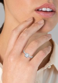 Anello con diamante taglio squadrato incastonato in argento, con dettagli intricati sulla banda, caratterizzato da un alone di diamanti più piccoli attorno alla pietra principale.