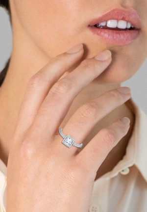 Anello con diamante taglio squadrato incastonato in argento, con dettagli intricati sulla banda, caratterizzato da un alone di diamanti più piccoli attorno alla pietra principale.