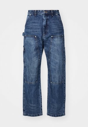 Mies pukeutuneena mustaan DSquared2-takkiin, jossa valkoinen "ICON"-teksti, keltainen DSquared2-t-paita, siniset leveälahkeiset farkut ja mustat kengät, seisoo sisätiloissa.