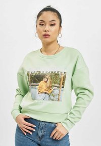 Lys grønn hettegenser med grafisk trykk av en kvinne utendørs på forsiden, med ribbestrikket krage og mansjetter, kombinert med høyt liv denim jeans.