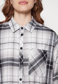 Camicia a quadri bianca e nera con colletto, dotata di taschino sul petto, bottoni neri e un tessuto morbido e dalla texture piacevole.