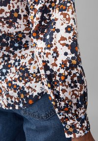 Blusa a maniche lunghe floreale con base bianca e motivi blu navy, marroni e arancioni, realizzata in tessuto leggero con polsini abbottonati.