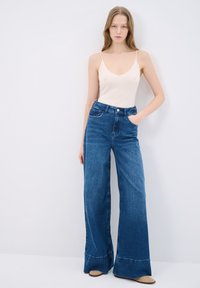 Jeans de tiro alto y pierna ancha en denim azul oscuro con bolsillos frontales y dobladillos a la altura del tobillo, combinados con una blusa de tirantes finos beige.