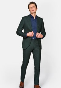 Ciemnozielony garnitur o dopasowanym kroju, z teksturowanej tkaniny, kontrastową granatową koszulą i brązowymi skórzanymi butami. Szczególne detale to klapy oraz guziki.