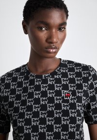 Μαύρο T-shirt με επαναλαμβανόμενο γκρι σχέδιο "PKNY", στρογγυλή λαιμόκοψη, κοντά μανίκια και ένα μικρό κεντημένο λογότυπο σε κόκκινο και λευκό.