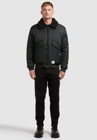 Giacca bomber nera con collo in ecopelliccia testurizzata, tasche frontali e polsini a costine. Indossata sopra pantaloni neri e stivali neri.