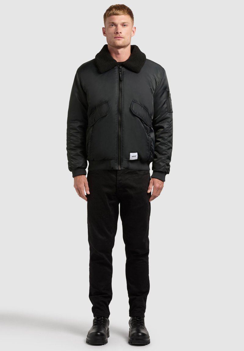 Giacca bomber nera con collo in ecopelliccia testurizzata, tasche frontali e polsini a costine. Indossata sopra pantaloni neri e stivali neri.