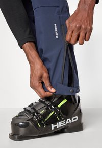 Ναυτικές σκιές παντελόνια από αδιάβροχο υλικό, με φερμουάρ κοντά στο κάτω μέρος. Ένα μαύρο σκι boot με πράσινες λεπτομέρειες είναι ορατό.