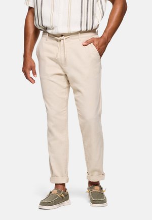 Hombre con pantalones beige de cordón con puños enrollados, camisa a rayas metida, y zapatos casuales verde oliva con suelas blancas.
