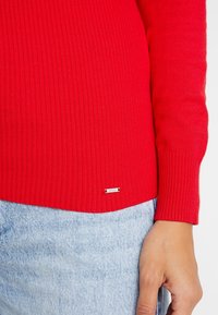 Hand in einer hellblauen Jeans neben einem roten gerippten Pulloverkanten mit kleinem silbernen rechteckigen Markenschild nahe dem Saum.