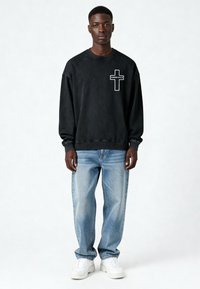 Jeune homme debout portant un sweat-shirt noir avec un motif croisé "KARMA KILLS", un jean bleu clair et des baskets blanches sur un fond uni.