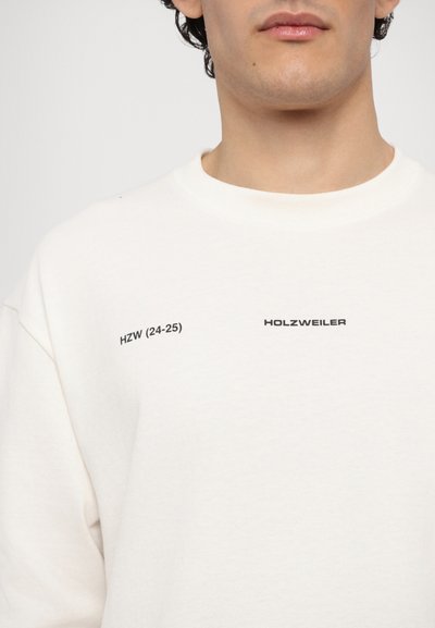 Svetřík v off-white barvě s kulatým výstřihem. Na přední straně je černý text "HZW (24-25)" a "HOLZWEILER". Má měkkou strukturu.